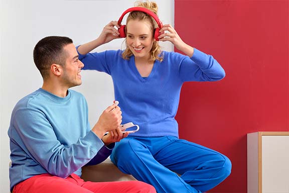 Zwei Personen sitzen auf dem Boden und interagieren miteinander. Eine Frau trägt rote Kopfhörer und lächelt, während ein Mann ein Audiogerät in der Hand hält.