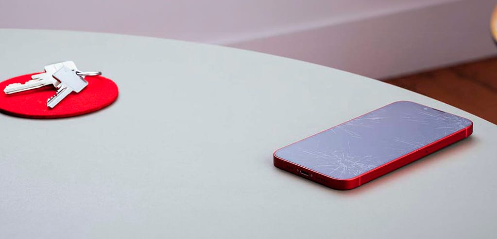 iPhone 12: Schutzfolie nötig? Rotes iPhone mit zerkratztem Display liegt auf einem Tisch neben einem Schlüsselbund auf einem roten Untersetzer
