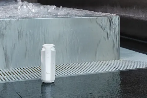  Ein weißer tragbarer Bluetooth-Lautsprecher steht auf einer nassen Oberfläche.