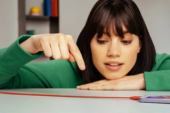 Person zeigt auf ein Flachband-Datenkabel, welches an ein Smartphone angeschlossen ist und auf einem Tisch liegt.