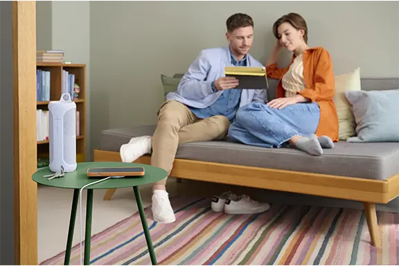 iPhone liegt auf einem induktiven Ladegerät und wird geladen. Im Hintergrund sitzen zwei Personen auf einem Sofa