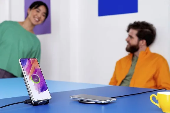 Smartphone auf einem Tisch mit kabelloser Ladestation und USB-Ladeoption, zwei Personen im Hintergrund in moderner Umgebung.