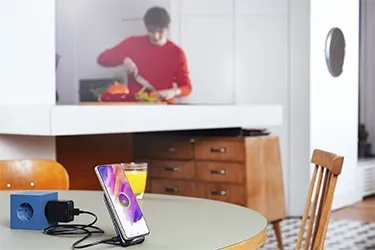 Smartphone auf einem Tisch mit Schnellladegerät, im Hintergrund eine Person beim Kochen in der Küche