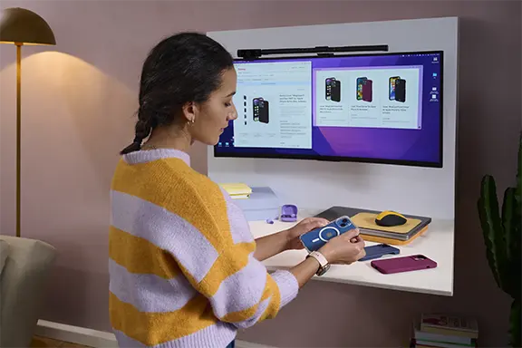 Person sitzt an einem Schreibtisch auf dem verschiedene Handyhüllen liegen. Auf dem Monitor sind verschiedene Produktseiten von Handyhüllen geöffnet.