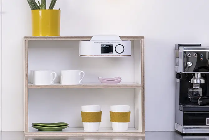 Weißer DAB+ Küchenradio ist an einem Küchenregal befestigt. Außerdem sind Tassen, Geschirr und eine Kaffeemaschine zu sehen.