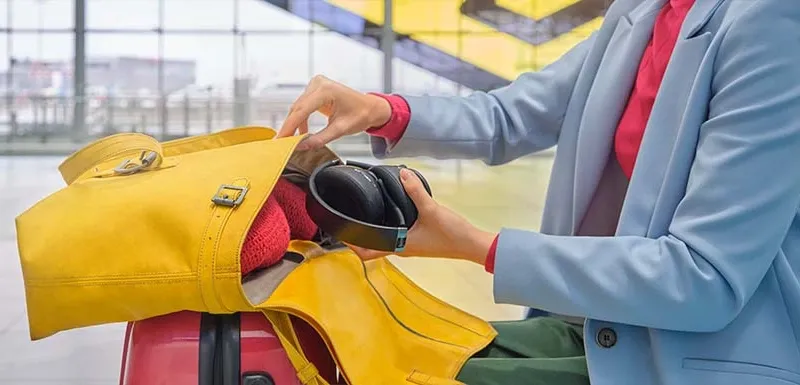 Bluetooth-Kopfhörer von Hama für Flugzeugreisen Person sitzt auf Wartebank im Flughafen und packt Bluetooth Over-Ear Kopfhörer in gelbe Tasche
