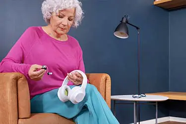 Ältere Frau mit Over-Ear-Kopfhörern und Hörgerät. Ältere Frau im pinken Oberteil und blauen Hosen hält weiße Over-Ear-Kopfhörer und ein Hörgerät in der Hand.