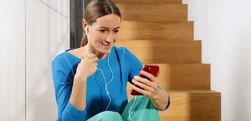 Frau mit Kopfhörern und Handy Frau mit blauem Oberteil sitzt auf einer Treppe, sie hat ein Handy in der Hand und Kopfhörer im Ohr