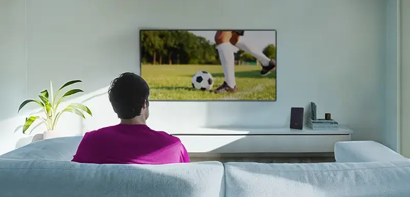 Person sitzt auf Sofa und schaut zum an der Wand hängenden Fernseher