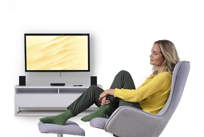 Frau in gelbem Pullover entspannt in einem modernen Wohnzimmer auf einem grauen Sessel, während sie auf einen Fernseher mit sandfarbenem Hintergrund blickt.