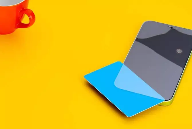Panzerglas wird mithilfe einer blauen Kreditkarte von einem Handy-Display entfernt, daneben steht eine rote Tasse