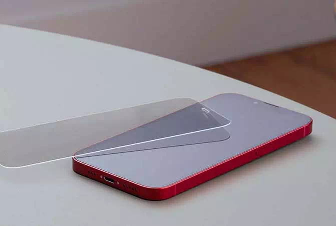 Ein rotes Smartphone liegt auf einer grauen Oberfläche, daneben ein transparentes Panzerglas für den Bildschirm von Hama, das leicht schräg über dem Display liegt
