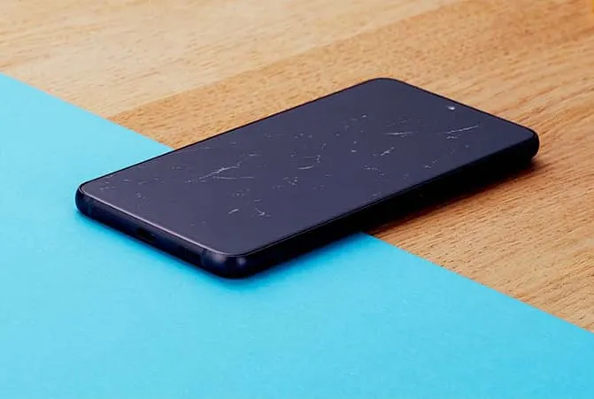 Ein Smartphone mit mehreren Kratzern auf dem Display, liegend auf einer Oberfläche, die zur Hälfte aus hellem Holz und zur Hälfte aus blauem Material besteht.