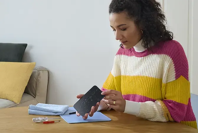 Eine Frau mit gestreiftem Pullover inspiziert den Bildschirm ihres Smartphones auf Kratzer, während sie an einem Tisch sitzt. Werkzeug zur Reparatur liegt daneben bereit