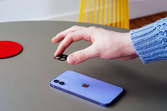 Anbringen eines Kameraschutzes auf einem iPhone Person installiert einen Hama Kameraschutz auf einem iPhone, das flach auf einem Tisch liegt