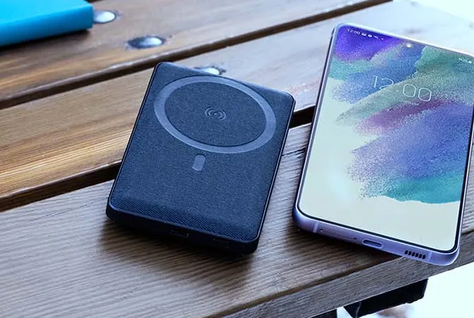 Eine schwarze Powerbank liegt neben einem Smartphone auf einem Holztisch im Außenbereich. Das Smartphone zeigt eine Uhrzeit auf dem Display an.