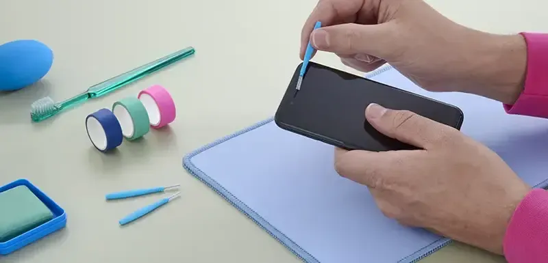 Person reinigt Lautsprecher eines iPhones mit einer kleinen Bürste. Daneben liegen weitere Hilfsmittel zur Reinigung.