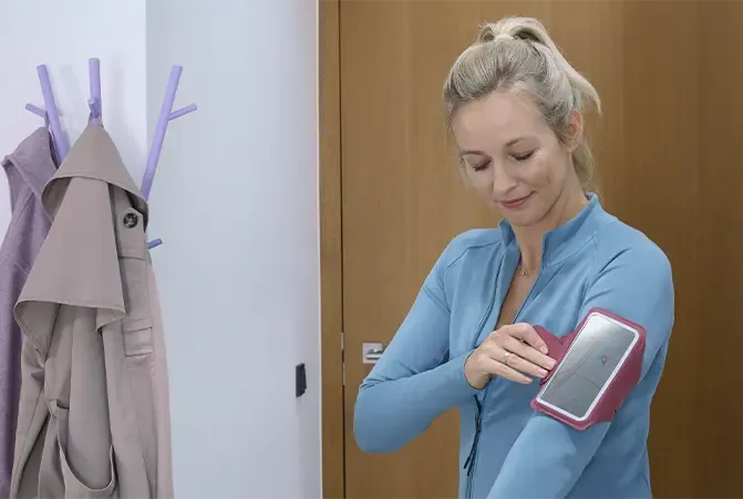 Frau steht im Flur und befestigt ihr Smartphone mit einem Hama Sportarmband am Arm. Im Hintergrund ist ein Garderobenständer und eine Tür zu sehen.