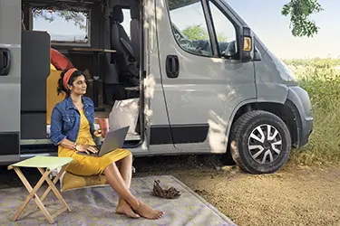 Frau arbeitet an einem Laptop und lädt ihn auf, während sie draußen neben einem Wohnmobil sitzt.