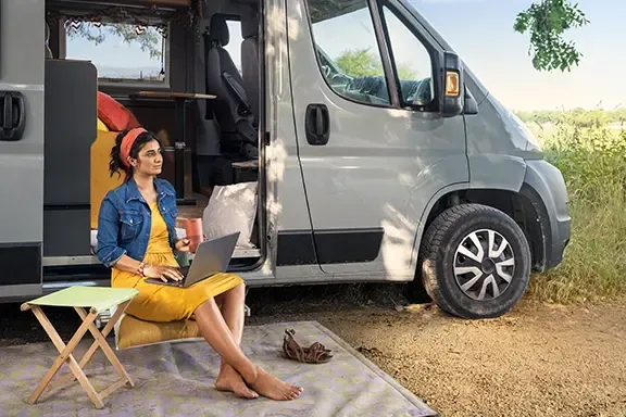 Frau arbeitet an einem Laptop und lädt ihn auf, während sie draußen neben einem Wohnmobil sitzt.