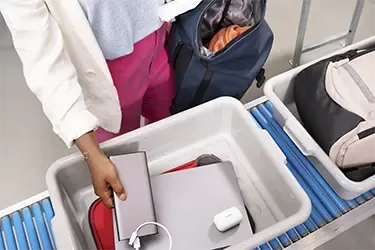 Laptop und Powerbank sicher durch die Flughafenkontrolle transportieren Person legt Laptop, Powerbank und Elektronik in eine Sicherheitsbox am Flughafen.