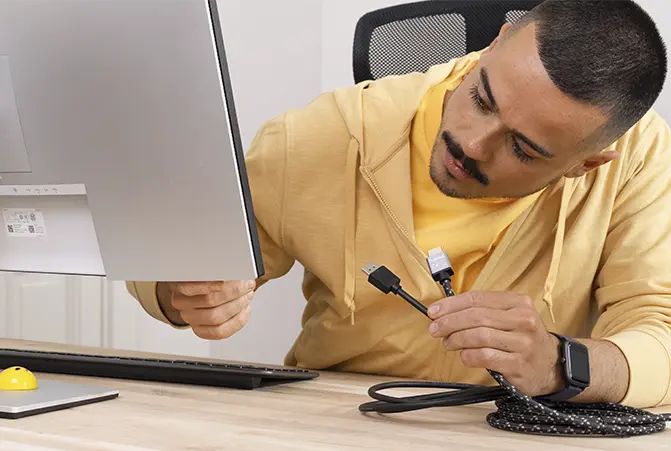 Person sitzt am Schreibtisch vor Monitor, hat Kabel mit DisplayPort & HDMI-Anschluss in der Hand und untersucht den Anschluss