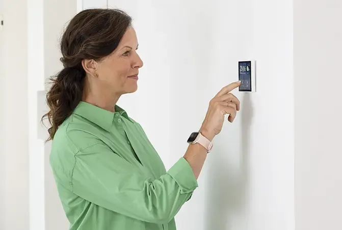 Frau in grüner Bluse bedient ein an der Wand montiertes Smart Home Thermostat