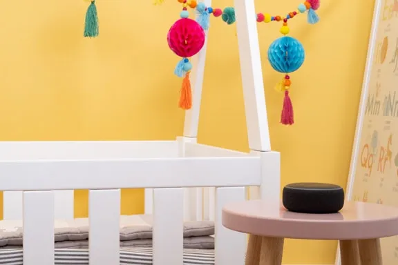 KInderbett steht vor gelber Wand mit Mobile, davor liegt Alexa auf dem Tisch und dient als Babyphone