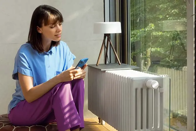 Person sitzt mit dem Smartphone in der Hand vor einem Heizkörper mit Smarthome-Thermostat.