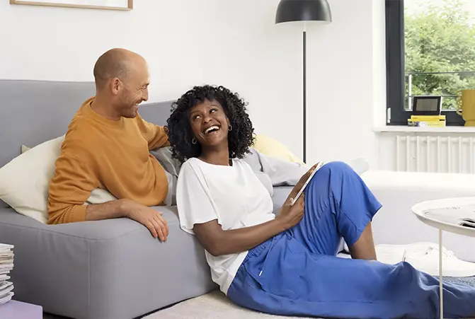 Zwei Personen sitzen lachend im Wohnzimmer und steuern die Smart-Home Geräte mit einem Tablet-PC.