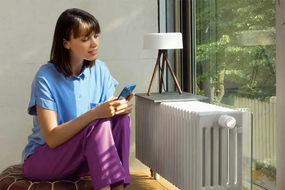 Person sitzt mit dem Smartphone in der Hand vor einem Heizkörper mit Smarthome-Thermostat.