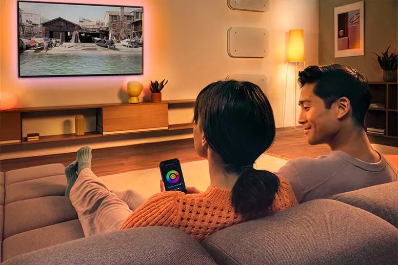 Zwei Personen sitzen auf der Couch und stellen via Smartphone gedimmtes Licht im Raum ein. 