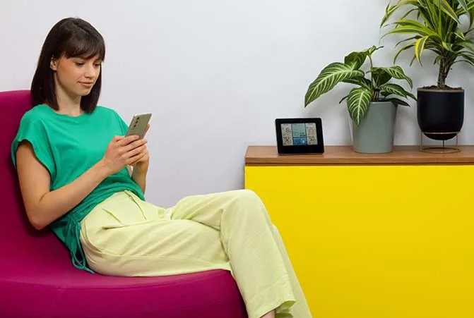 Eine Frau schaut auf ihr Smartphone. Neben ihr steht eine Wetterstation auf einem Sideboard.