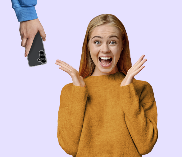 Eine junge Frau in orangem Pullover blickt überrascht und gut gelaunt in die Kamera. Aus der oberen linken Ecke hält eine Hand ein Samsung-Handy nach unten.